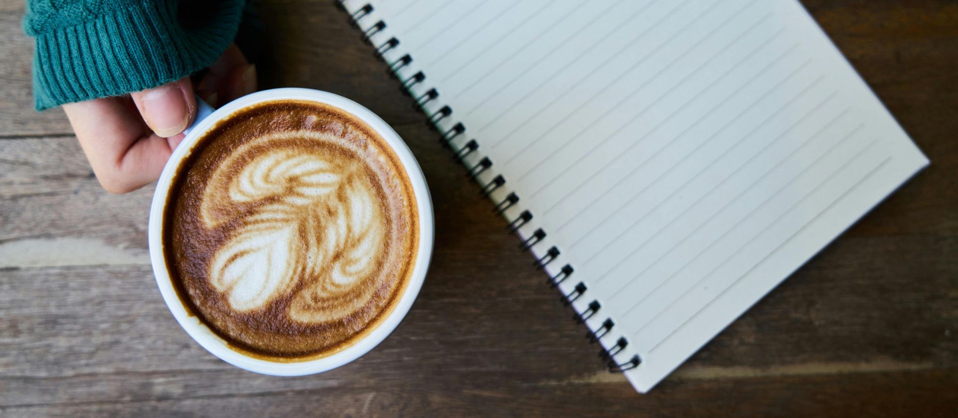 Top view of a latte with latte art next to a blank notebook on a wooden table.